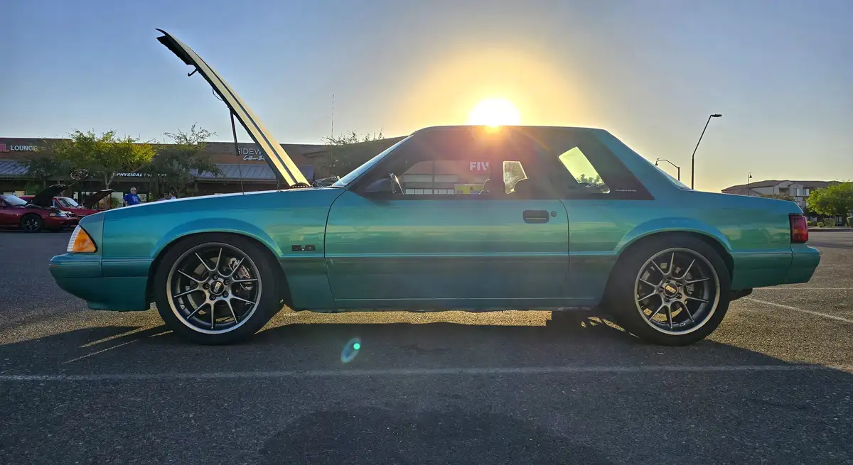 A teal Ford Mustang with its hood open is parked in a lot during sunset, with the sun directly behind it. The car has…