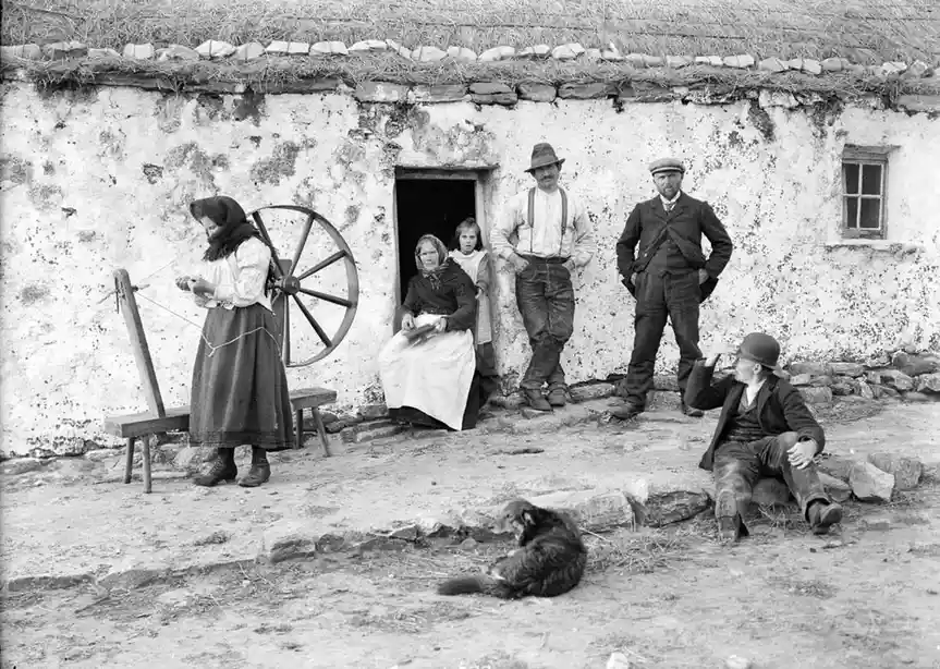 A vintage black-and-white photo shows a family of six posing outside a thatched-roof cottage. A woman spins wool at a…