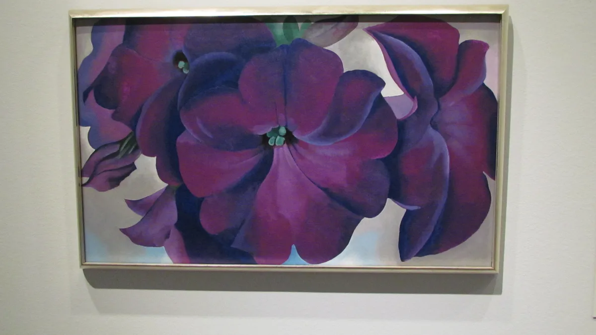 A framed painting of stylized purple flowers with thick, rounded petals and green centers, hanging on a white wall. The…