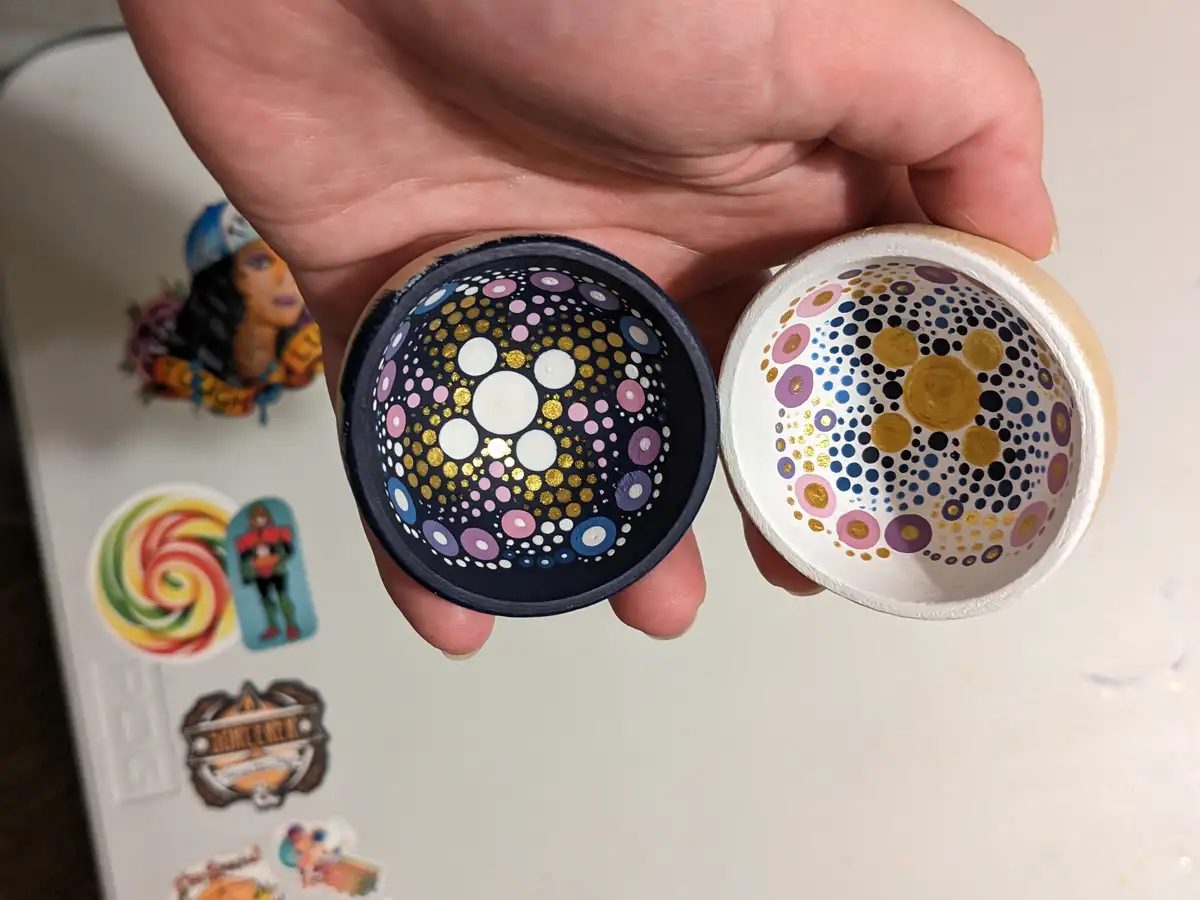 A hand holds two halves of a painted, spherical object, revealing intricate, colorful dot patterns on the inside resembling…