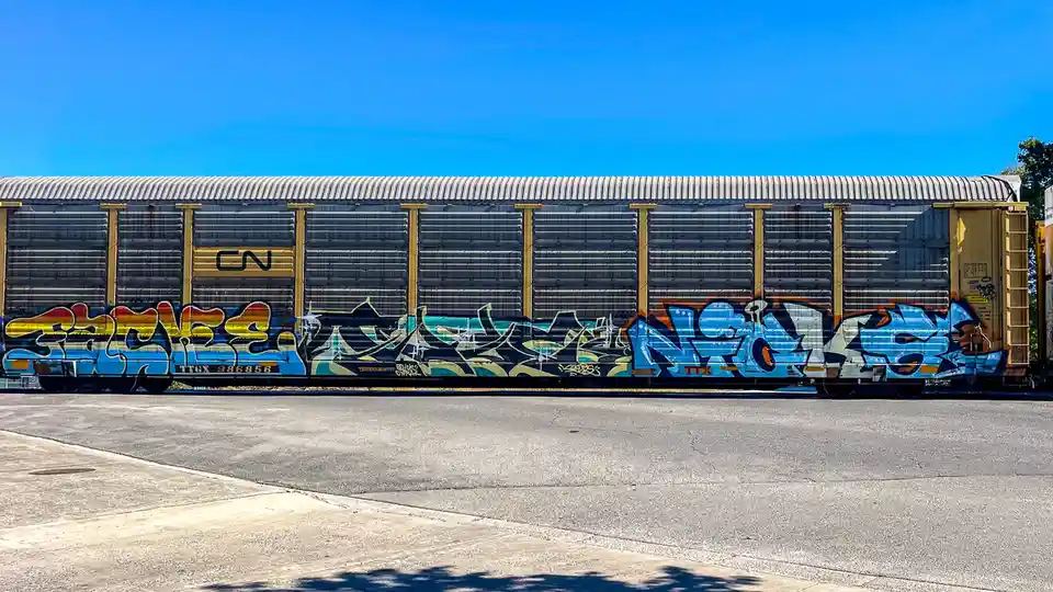 A side view of a long, silver CN freight train car covered in colorful graffiti under a clear blue sky. The overlaid text…