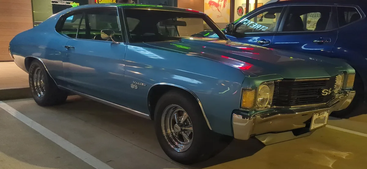 A shiny blue 1970s Chevrolet Malibu SS muscle car is parked in a lot at night, its chrome details and classic design…