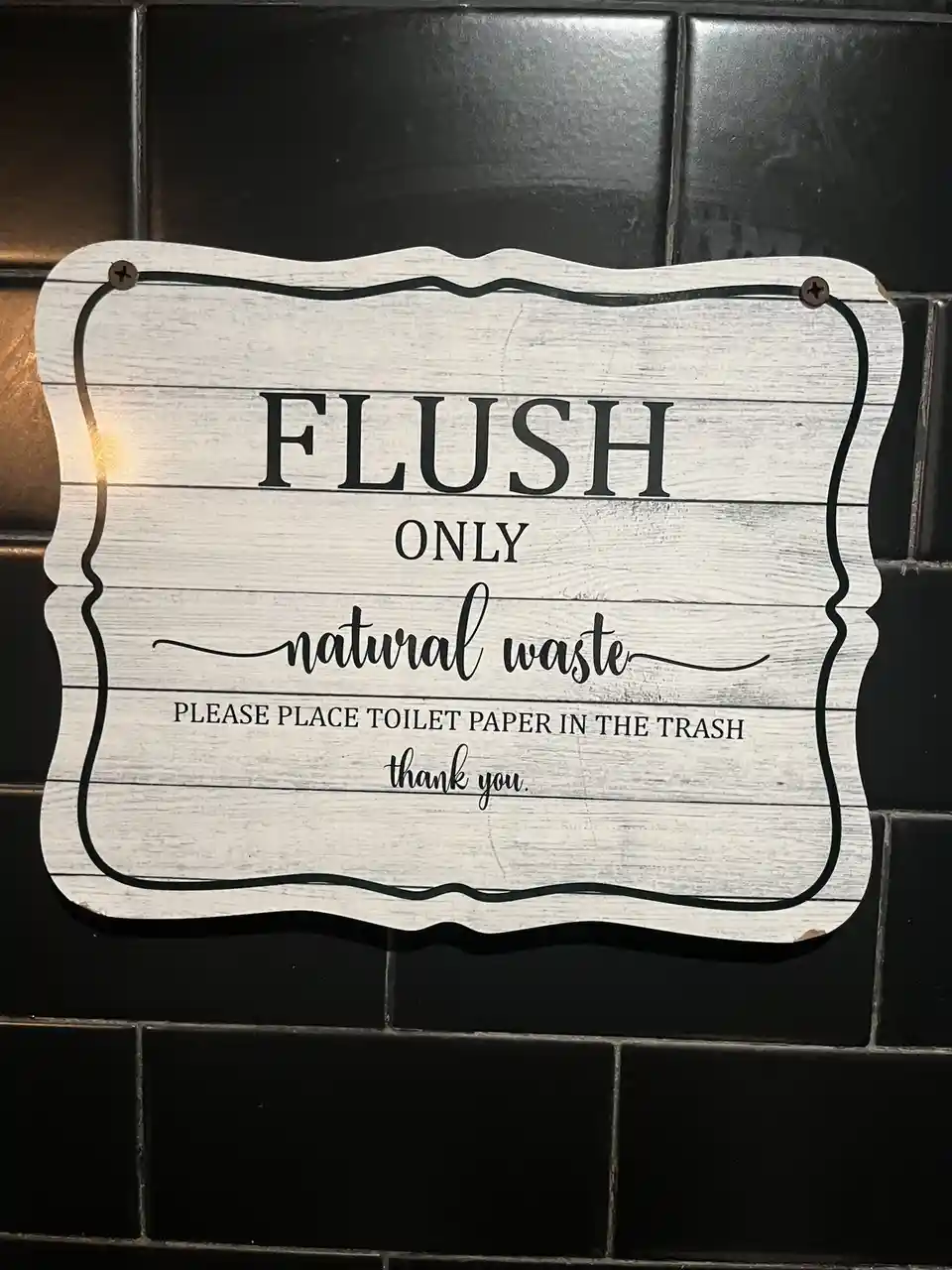 A white, weathered wooden sign with black text is mounted on a black tiled wall. The sign reads 'FLUSH ONLY natural waste'…