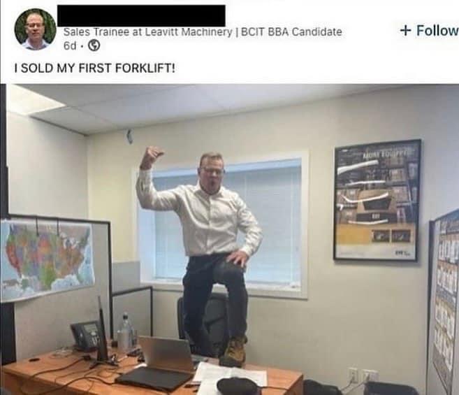 A man in a white shirt and glasses is standing on his office chair with one fist raised in triumph, celebrating in an office…