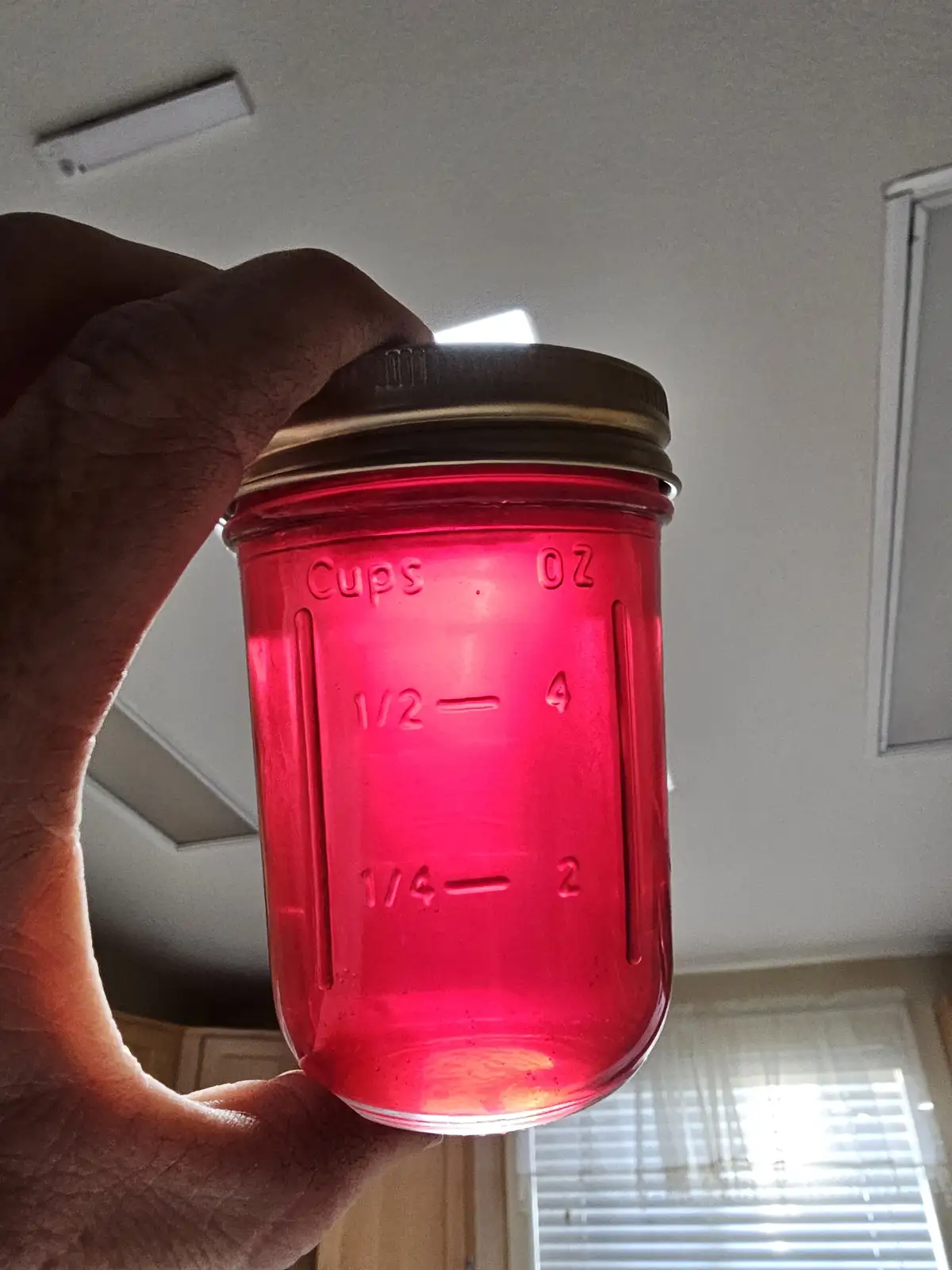 A hand holds a red-tinted glass mason jar with measurement markings, filled with a red liquid, against a kitchen background…