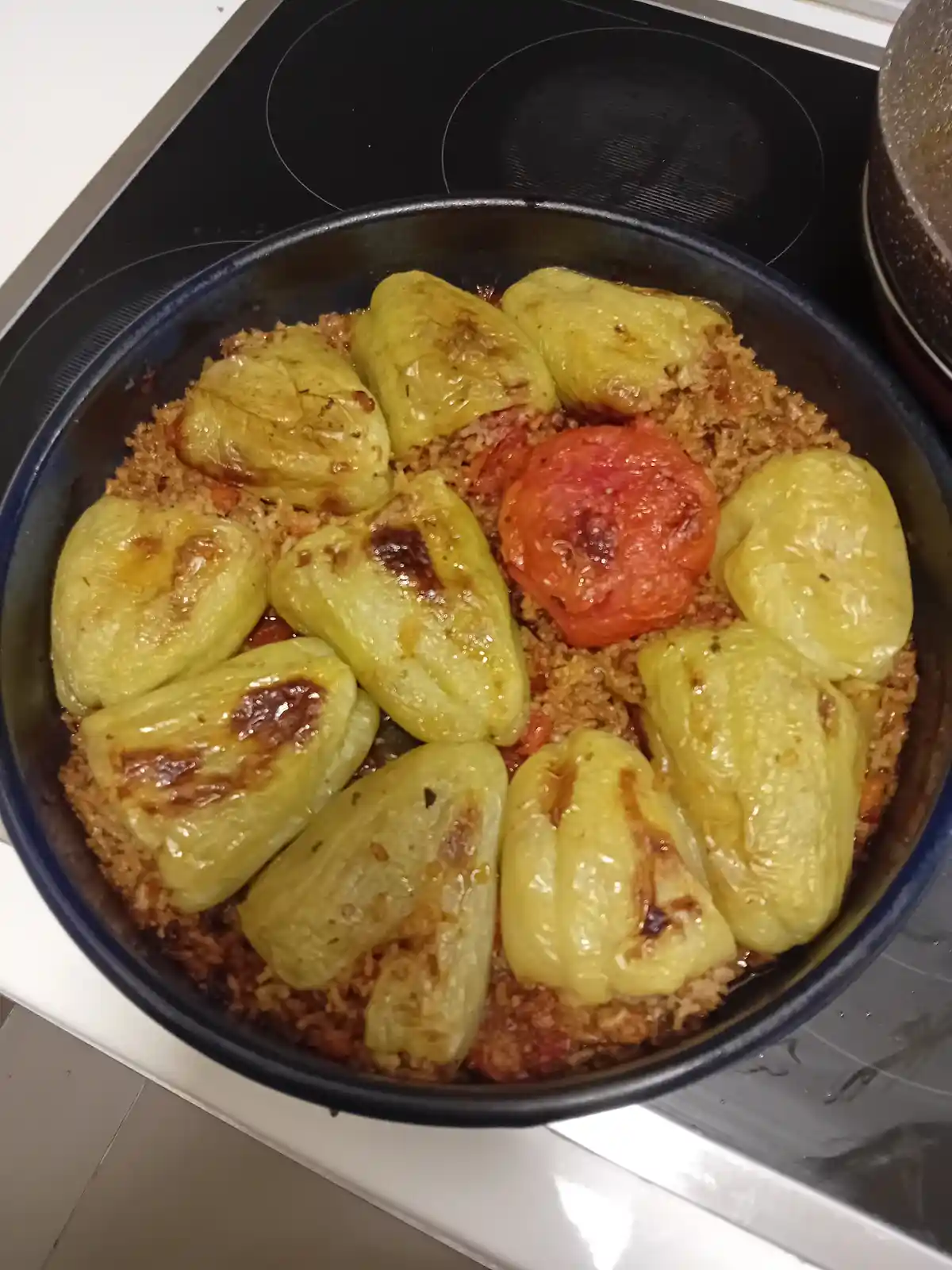 A round black baking dish sits on a stovetop, filled with a rice-based casserole topped with ten roasted yellow bell peppers…