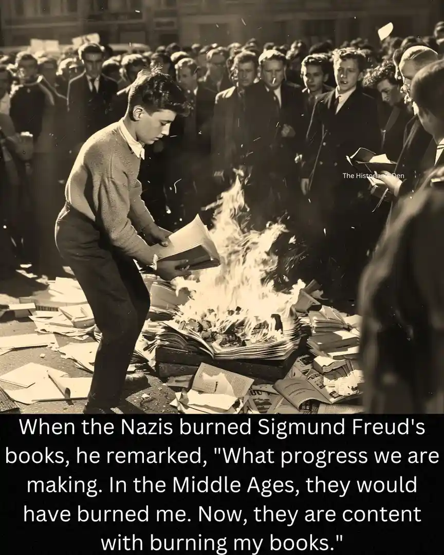 A black-and-white historical photograph shows a young boy in a sweater and trousers throwing books into a large bonfire…