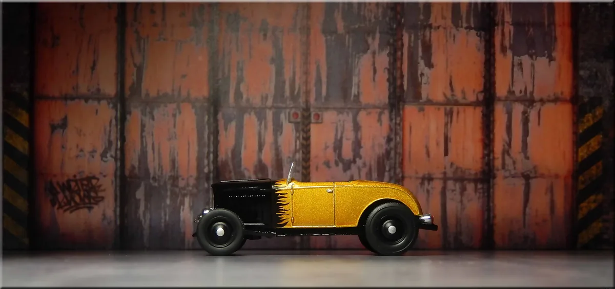 A shiny gold and black vintage hot rod toy car with flame decals is centered on a gray floor against a rusted, orange-brown…