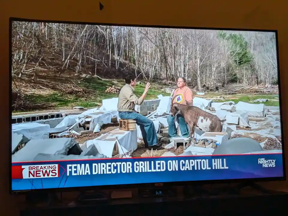 A television screen shows a news broadcast with a breaking news banner reading 'FEMA DIRECTOR GRILLED ON CAPITOL HILL'. The…