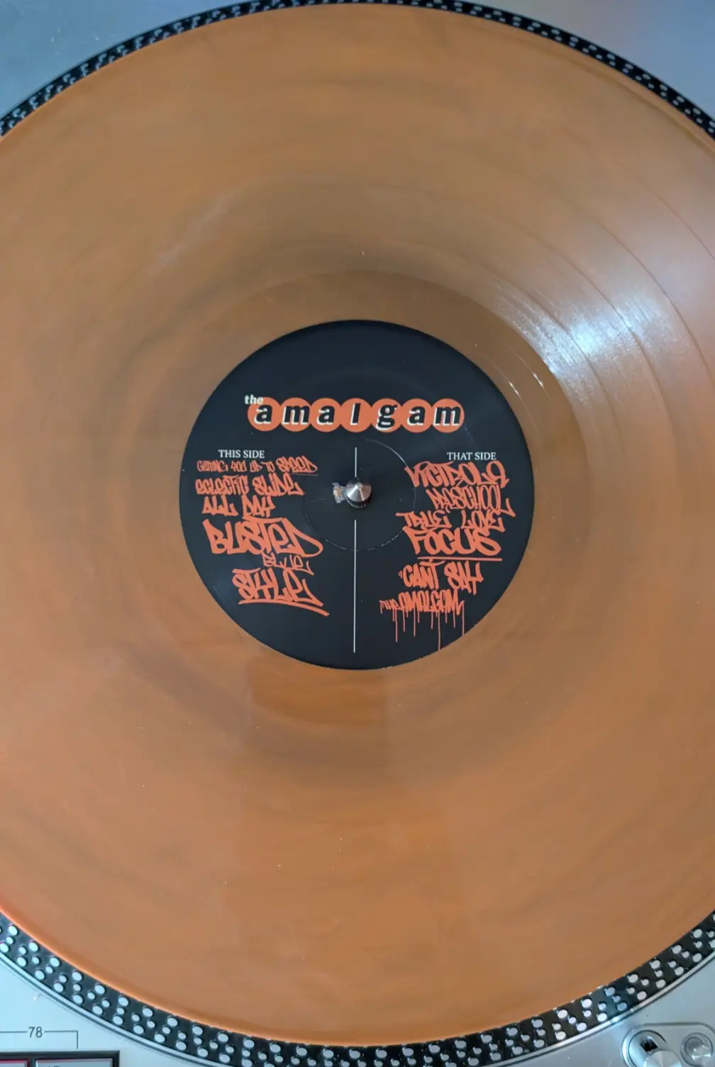 A close-up of a translucent orange vinyl record on a turntable, showing its black center label. The label has orange…