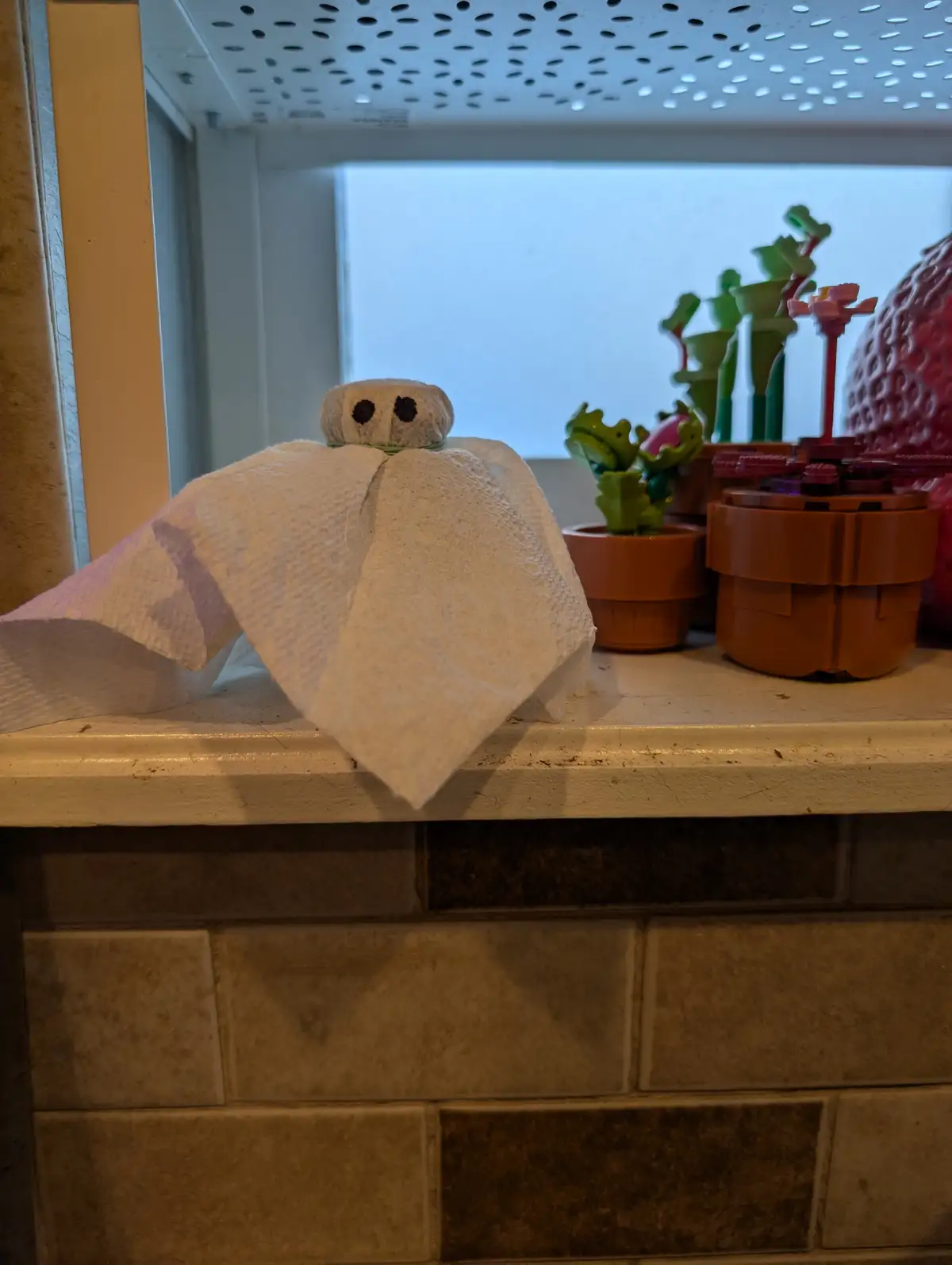 A paper towel is draped over a small object to resemble a ghost, with two black dots for eyes, sitting on a tiled windowsill…