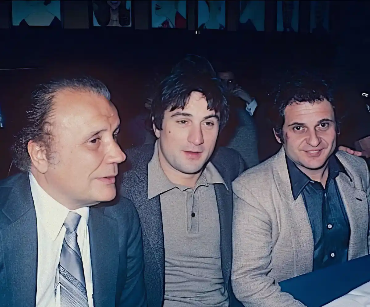 three men are seated side by side in a dimly lit room; the man on the left in a suit and tie looks toward the center, the…