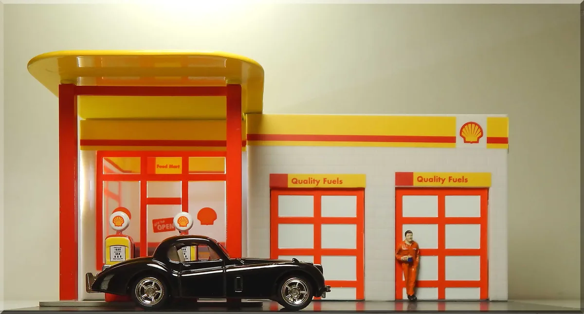 A miniature model of a Shell gas station with a black classic car parked at a pump, a small figurine of a worker in an…