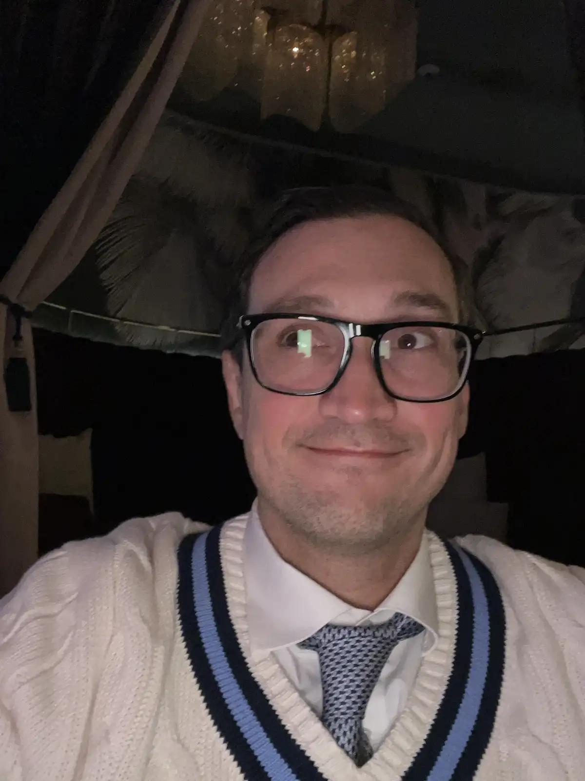 A man wearing glasses, a white collared shirt, a patterned tie, and a cream-colored V-neck sweater with blue and navy…