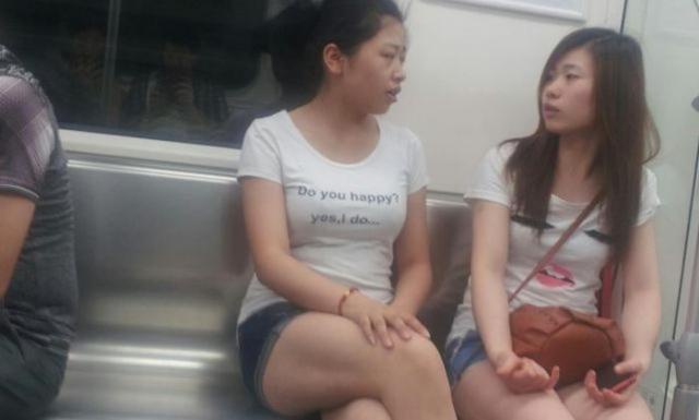 Two young women sit on a subway bench; the woman on the left wears a white t-shirt with the text 'Do you happy? yes, i…