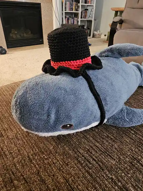 A large, blue, plush whale toy lies on its side on a brown carpet in a living room, wearing a black crocheted top hat with a…