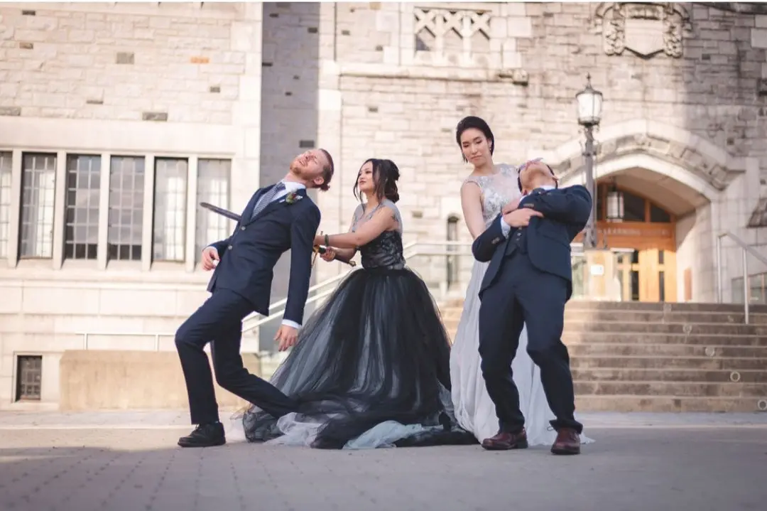 Four people dressed in wedding attire pose dramatically outdoors in front of a stone building. Two men in suits lean back…