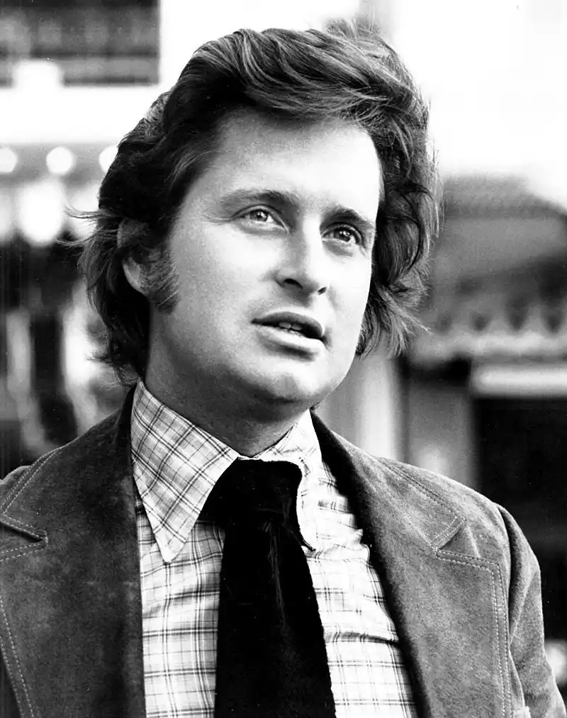 A black-and-white photo of a man with wavy hair, wearing a plaid shirt and dark tie under a jacket, looking upward with a…