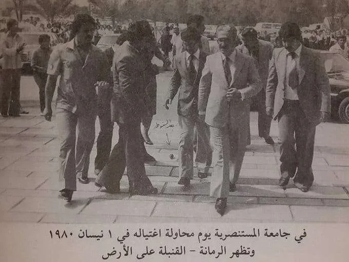 A black-and-white photo shows a group of men in suits walking on a paved area, one man in the center appears to be walking…