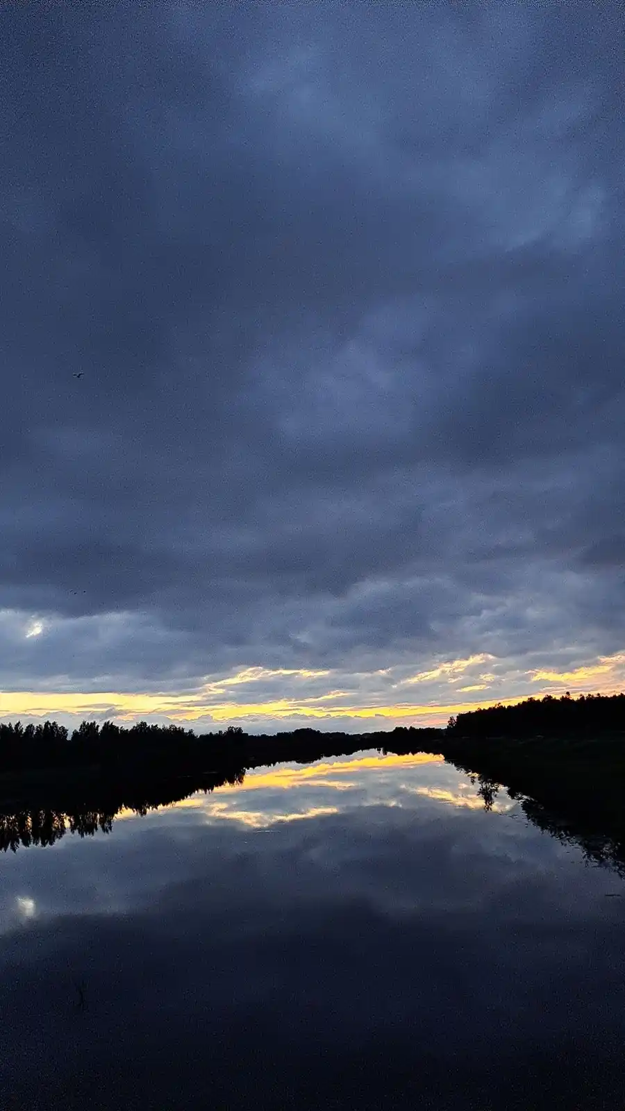 A serene landscape photo shows a calm body of water reflecting a dramatic sky at dusk or dawn, with dark clouds overhead and…