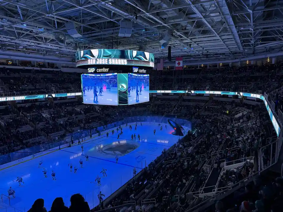 A wide-angle view of the SAP Center arena during a San Jose Sharks hockey game, showing players on the ice, a large central…