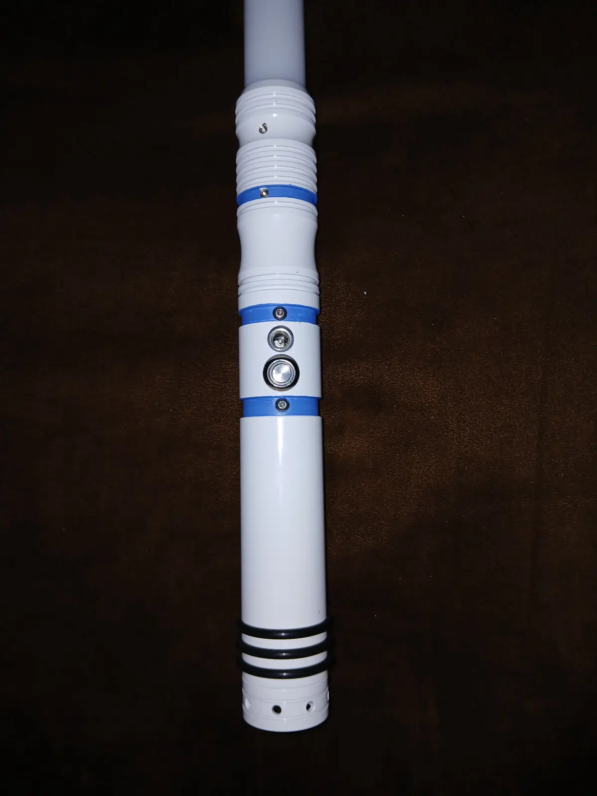 A close-up photo shows the hilt of a white lightsaber replica with blue and black accents, resting on a dark brown textured…