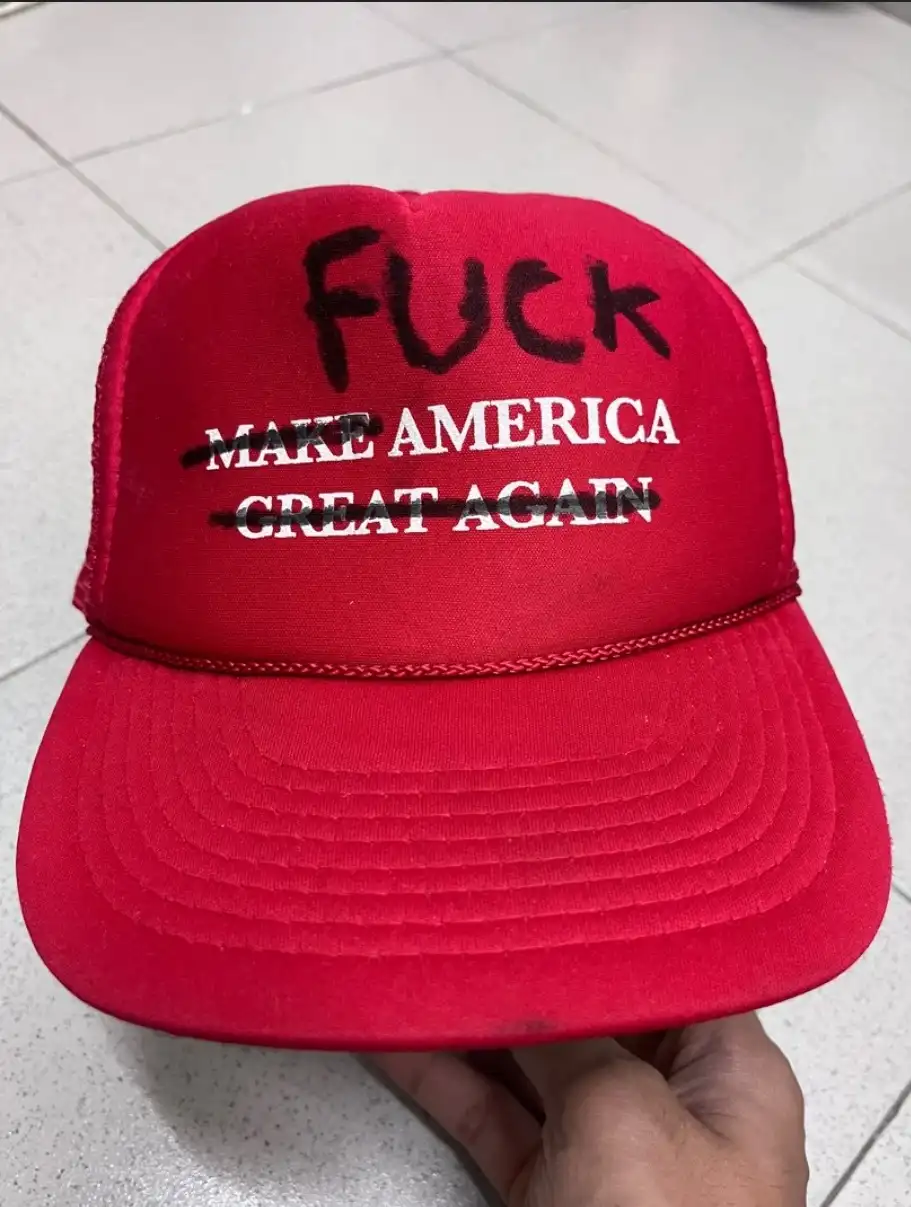 A red trucker hat with the phrase 'MAKE AMERICA GREAT AGAIN' printed in white, with both lines crossed out in black marker…