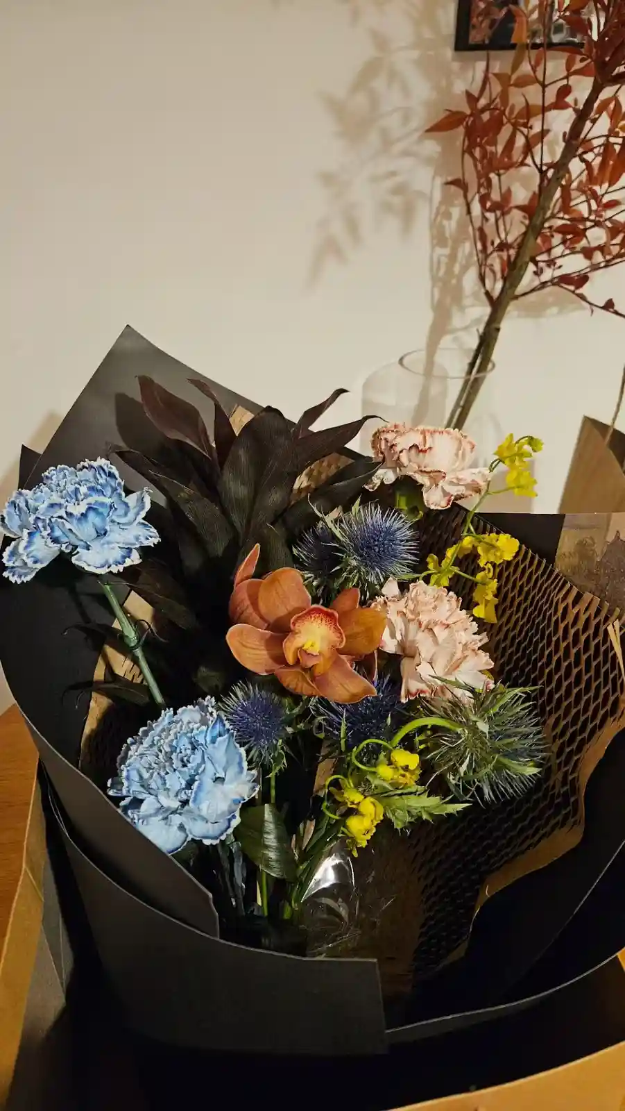 A close-up photo of a colorful, artfully arranged bouquet wrapped in black paper, featuring blue carnations, an orange…