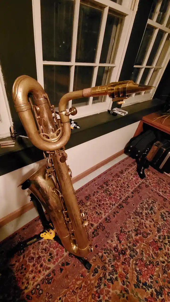 A brass saxophone is propped up on a stand indoors, positioned in front of a window with multiple panes. The instrument's…