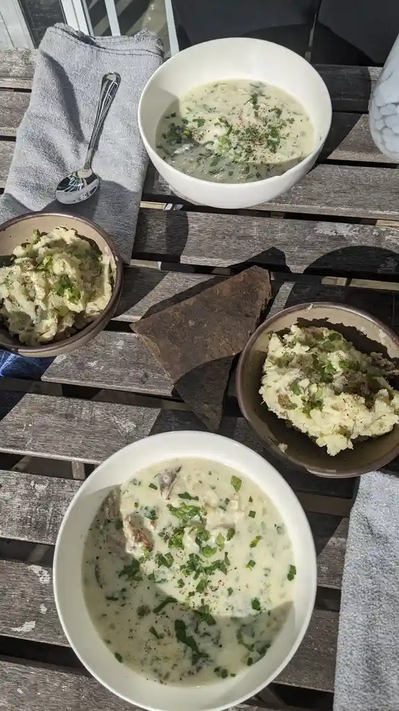 An overhead shot of a wooden table set with two bowls of creamy soup garnished with green herbs and black pepper, two bowls…