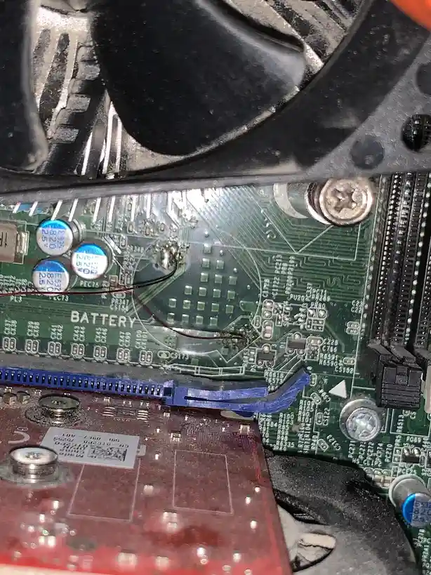 A close-up photo of a green computer motherboard with a labeled 'BATTERY' section, showing a small, round, silver CMOS…