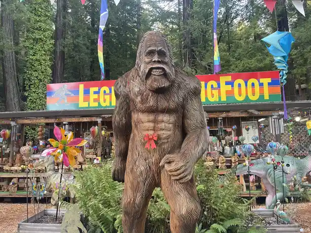 A large wooden statue of Bigfoot stands in front of a colorful shop with a sign reading 'LEGEND BIGFOOT'. The statue wears a…