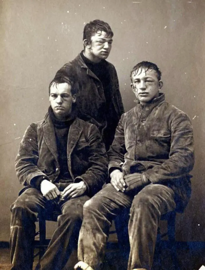 A sepia-toned vintage photograph shows three young men in worn, dark work clothes posing stiffly. The man standing behind…