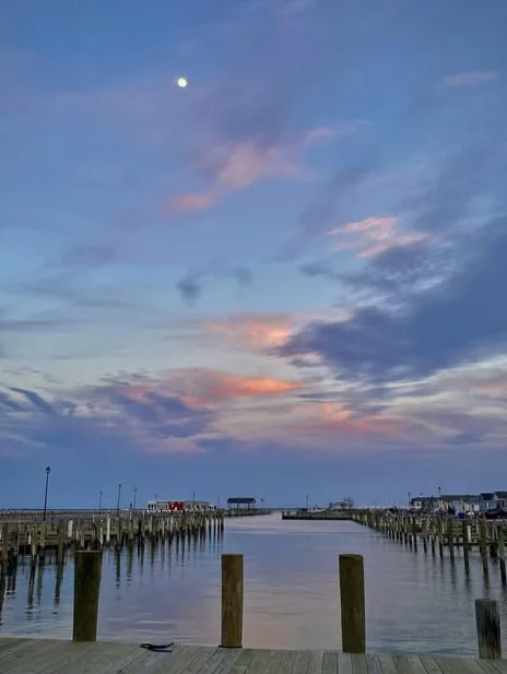 A serene twilight photo of a wooden pier extending over calm water, with a moon visible in the sky and soft pink and blue…