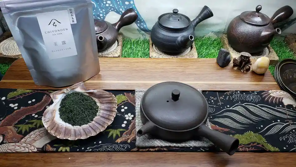 A serene Japanese tea setup features a bag of 'CHIYONOEN TEA FARM' matcha labeled '玉露', a shell holding green tea leaves…
