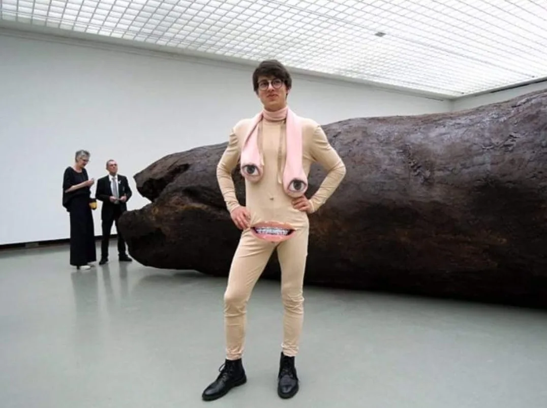 A person in a beige bodysuit with large cartoon eyes on a pink scarf and a mouth with braces at the waist stands confidently…