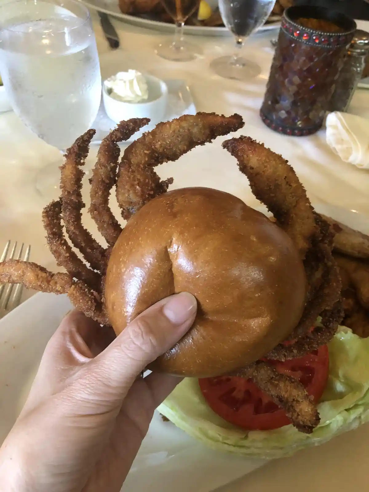 a hand holds a burger bun with fried, crab-leg-shaped protrusions resembling crab legs, sitting atop lettuce and tomato on a…