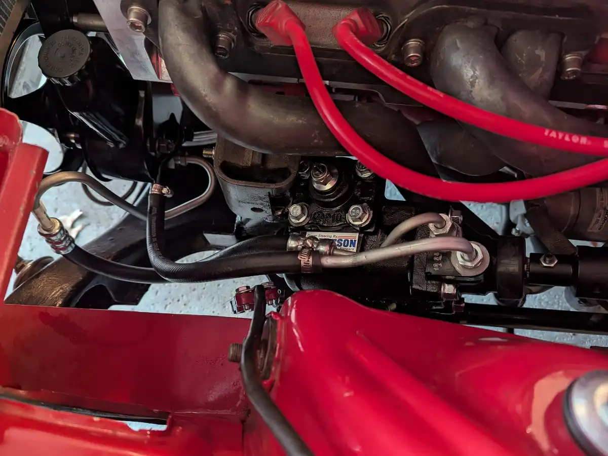 A close-up photo of a car's engine bay showing red and black hoses, bolts, and mechanical parts. A small sticker on the…