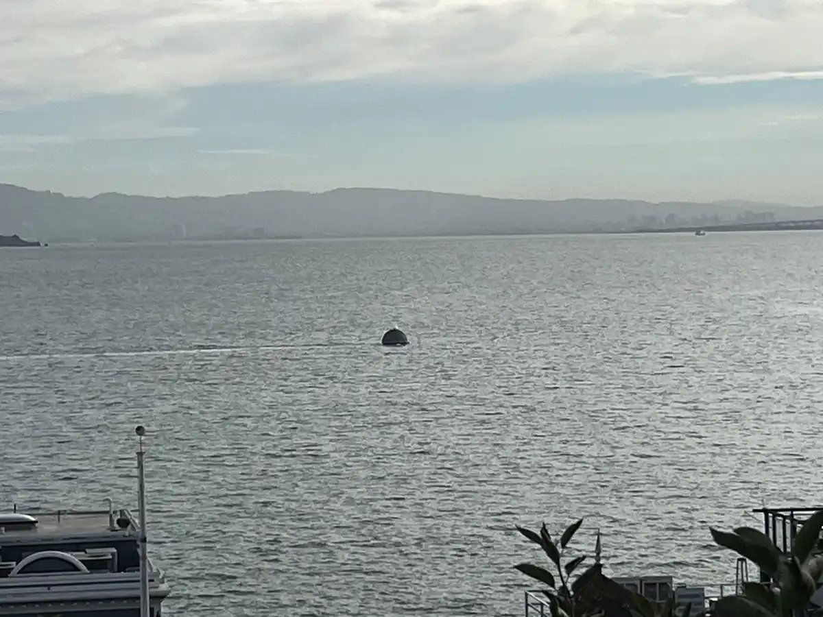 A wide shot of a calm body of water with distant hills and a city skyline under a cloudy sky. A small, dark, dome-shaped…