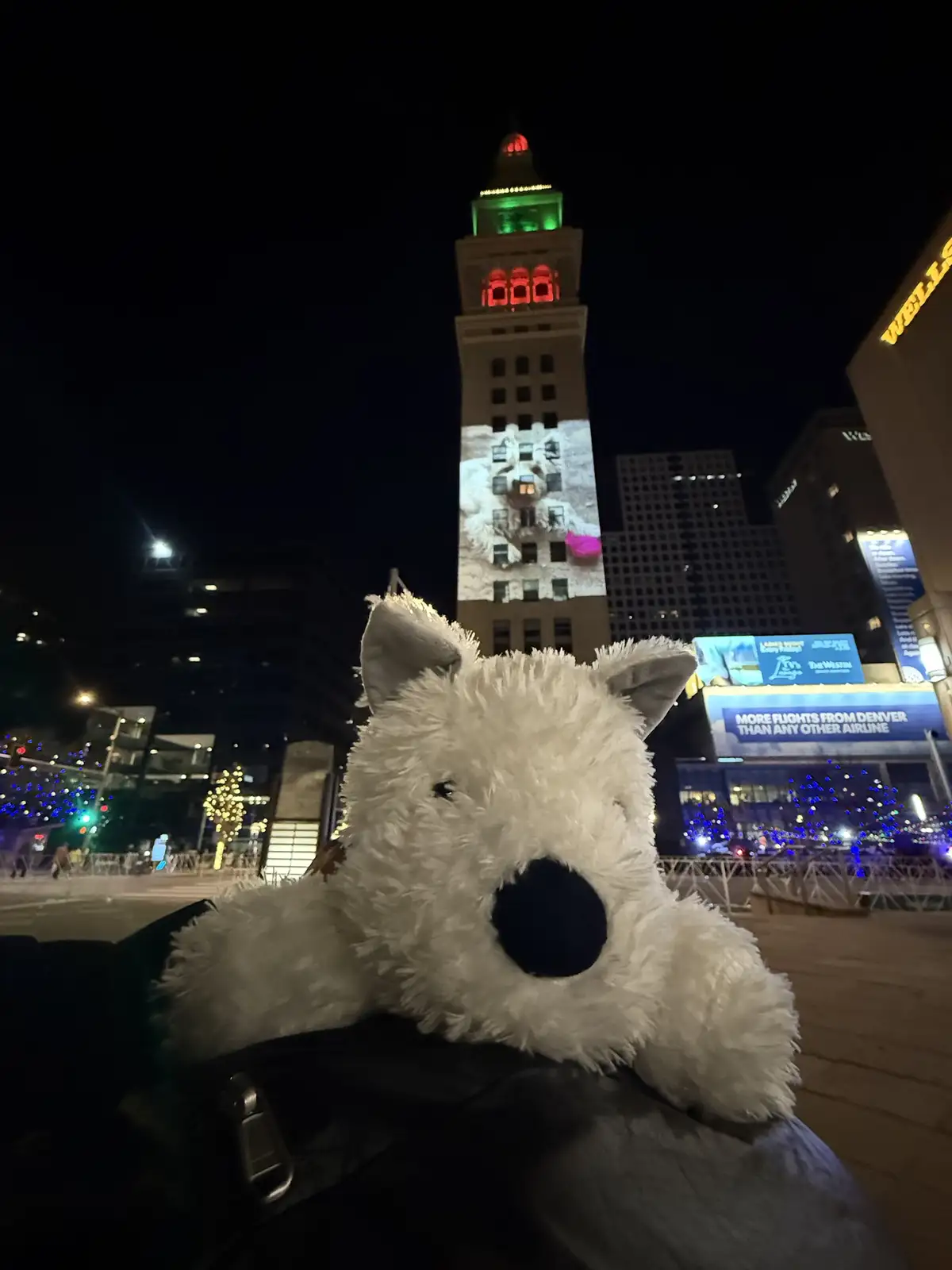 A white, fluffy stuffed dog toy is in the foreground, resting on a dark jacket, with a city skyline at night behind it. The…