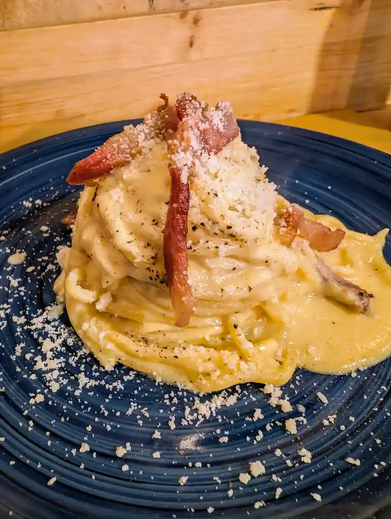 A close-up photo shows a tall, creamy pasta dish on a dark blue plate, topped with crispy bacon strips and grated cheese…