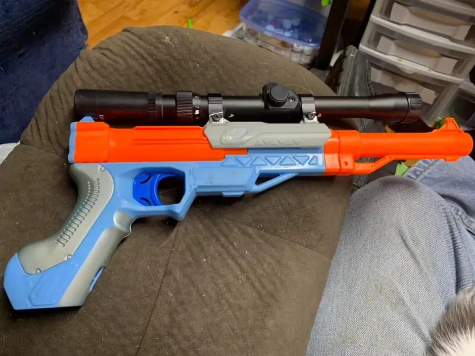 A blue and orange Nerf-style toy gun with a black scope mounted on top, resting on a person's lap. The person is wearing…