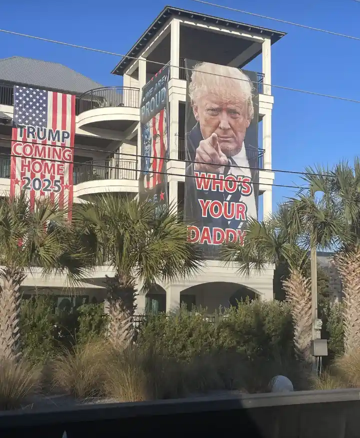 A building facade displays large political banners: one reads 'TRUMP COMING HOME 2025' with an American flag motif, and…