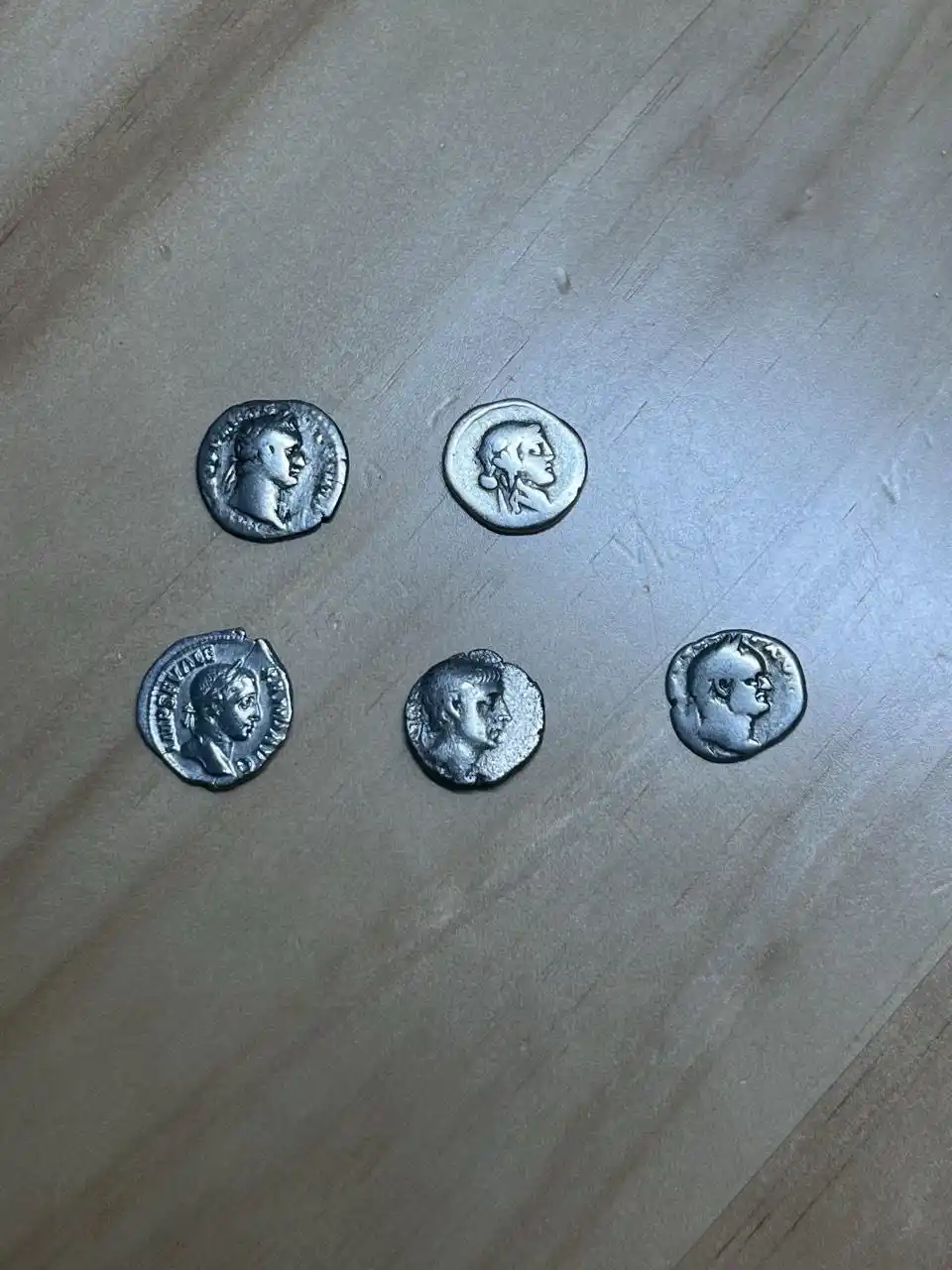 Five ancient Roman silver coins, each showing a profile bust of a ruler, are arranged on a light-colored wooden surface. The…