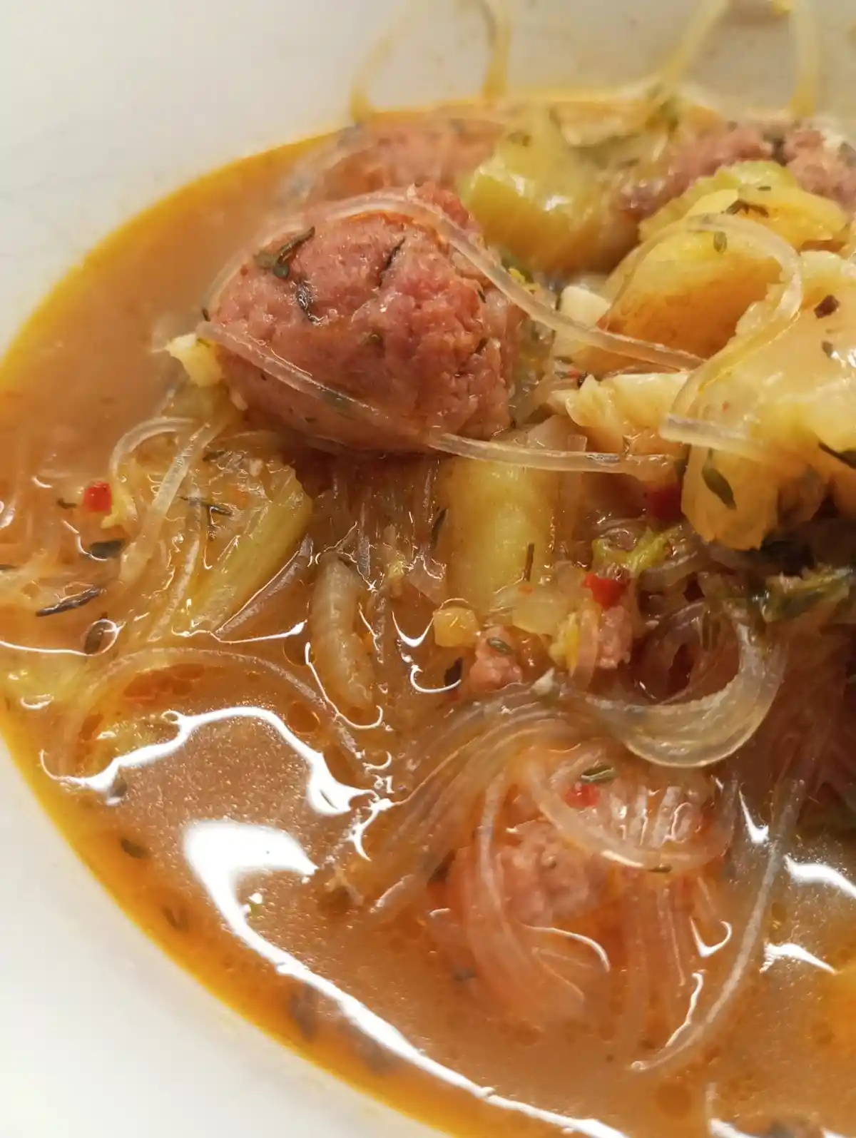 A close-up photo of a bowl of soup containing meatballs, translucent noodles, chopped vegetables, and herbs in a brown…
