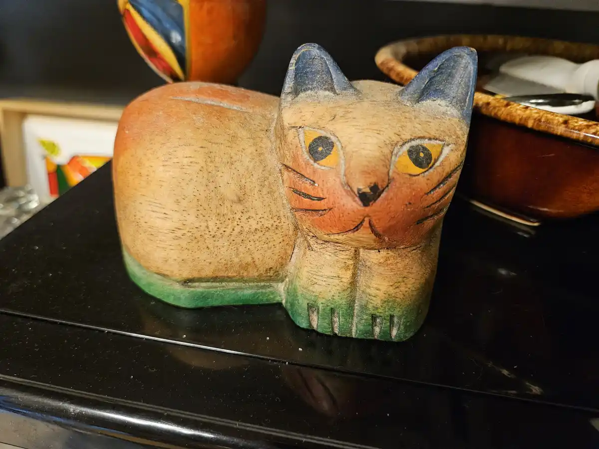 A wooden, hand-painted cat statue with blue ears, orange eyes, and green paws sits on a black surface. The overlaid text…