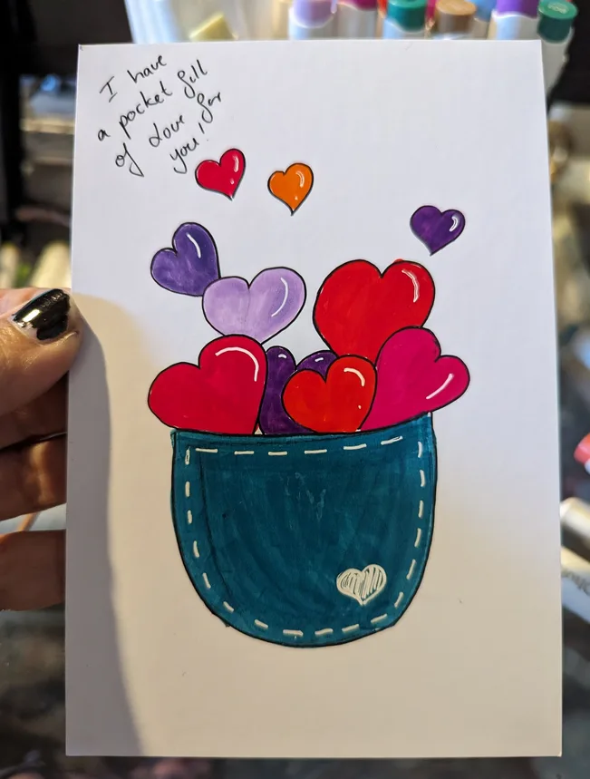 A hand holds a handmade card showing a blue denim pocket overflowing with colorful hearts. The handwritten text above reads…