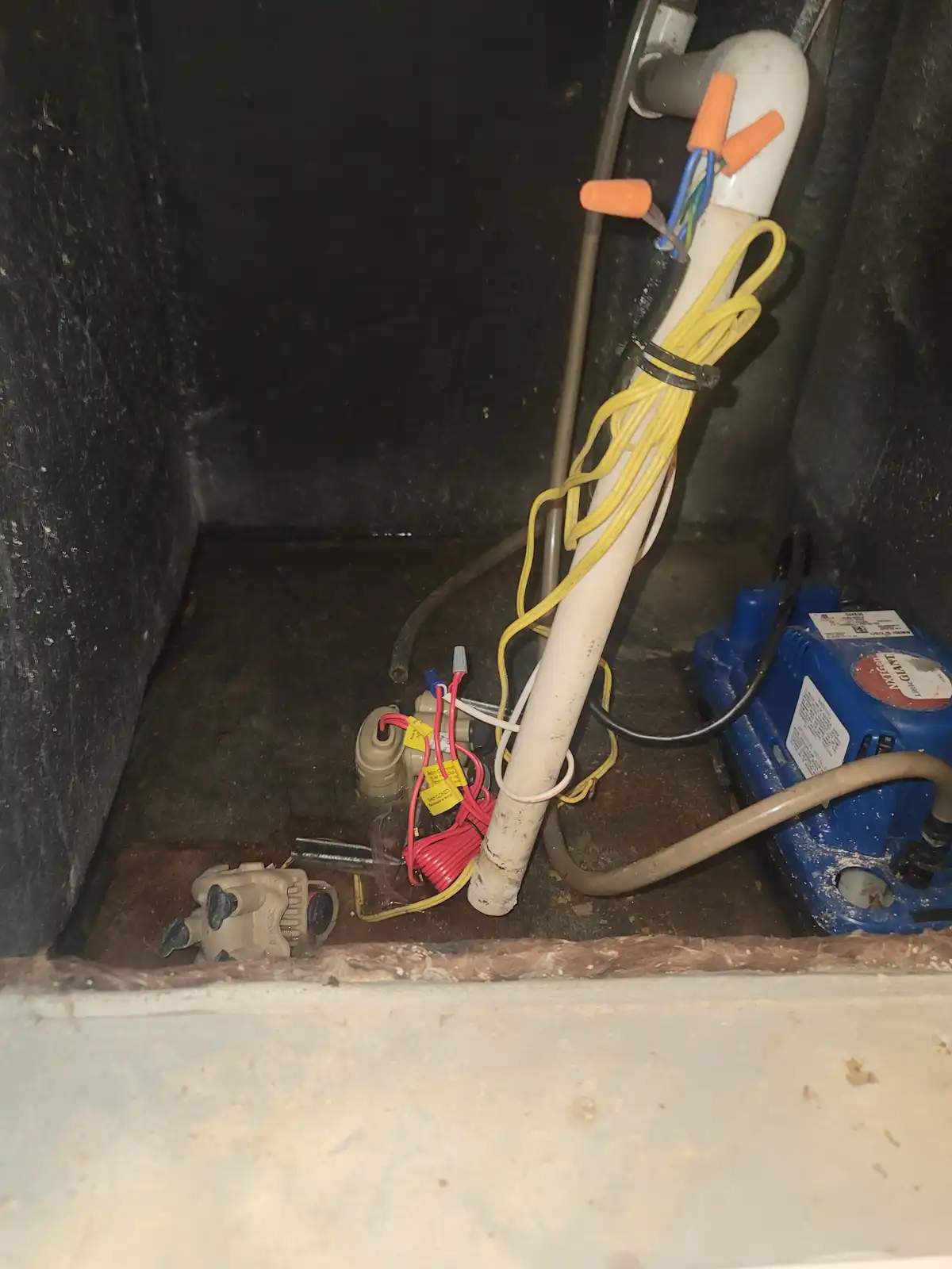 A cluttered, dimly lit utility space shows a white PVC pipe with orange caps and tangled yellow, red, and black wires. A…