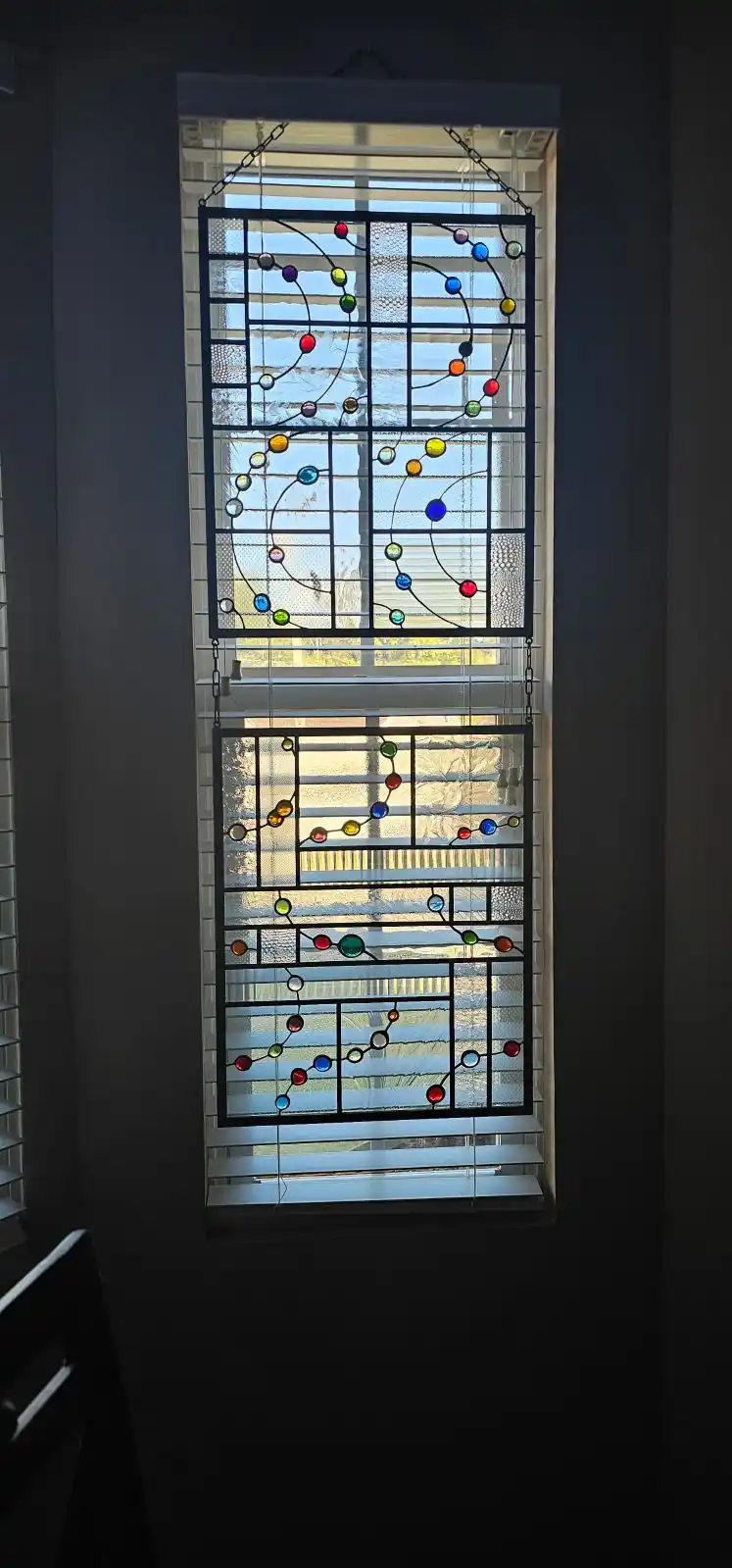 A stained glass window hanging in front of a window with blinds, featuring colorful circular glass pieces connected by lines…