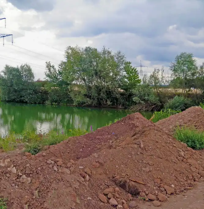 A photo of two large piles of reddish-brown dirt in the foreground, with a calm greenish river and trees behind them under a…